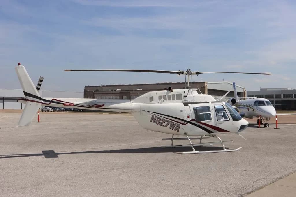 A white Bell 206 helicopter with N827WA registration on the side, parked on an airfield with a jet in the background.