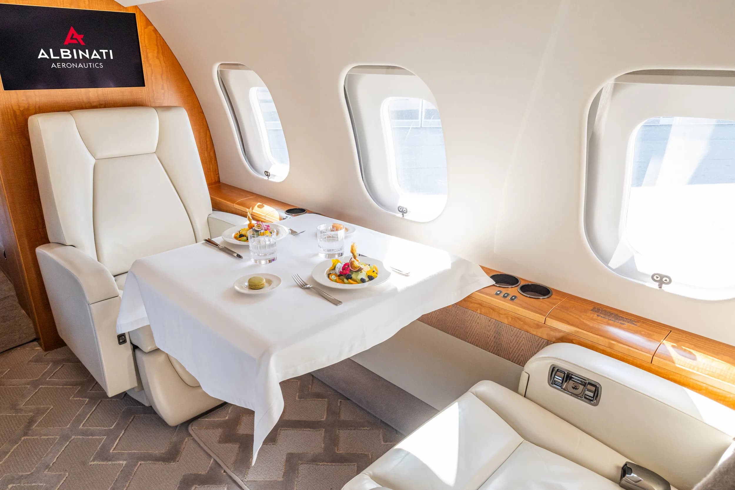 A dining table set with fruit and drinks in a private jet cabin, with an "ALBINATI AERONAUTICS" screen visible.