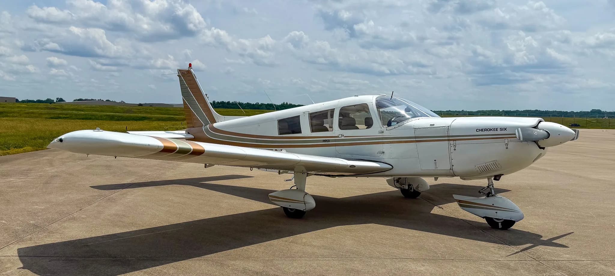 A front-right view of a white Piper Cherokee Six aircraft with brown and green stripes on an airfield under a cloudy sky.