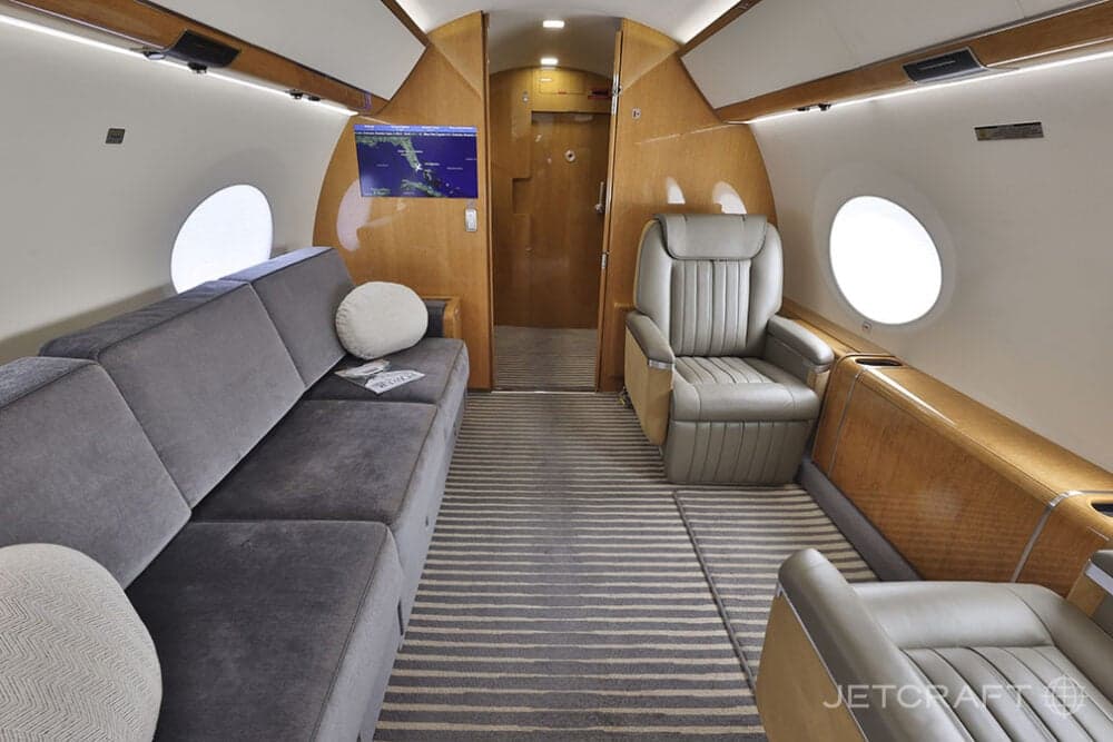 A view of the business jet cabin with a grey sofa, a light brown leather seat, and a wall-mounted screen displaying a map.