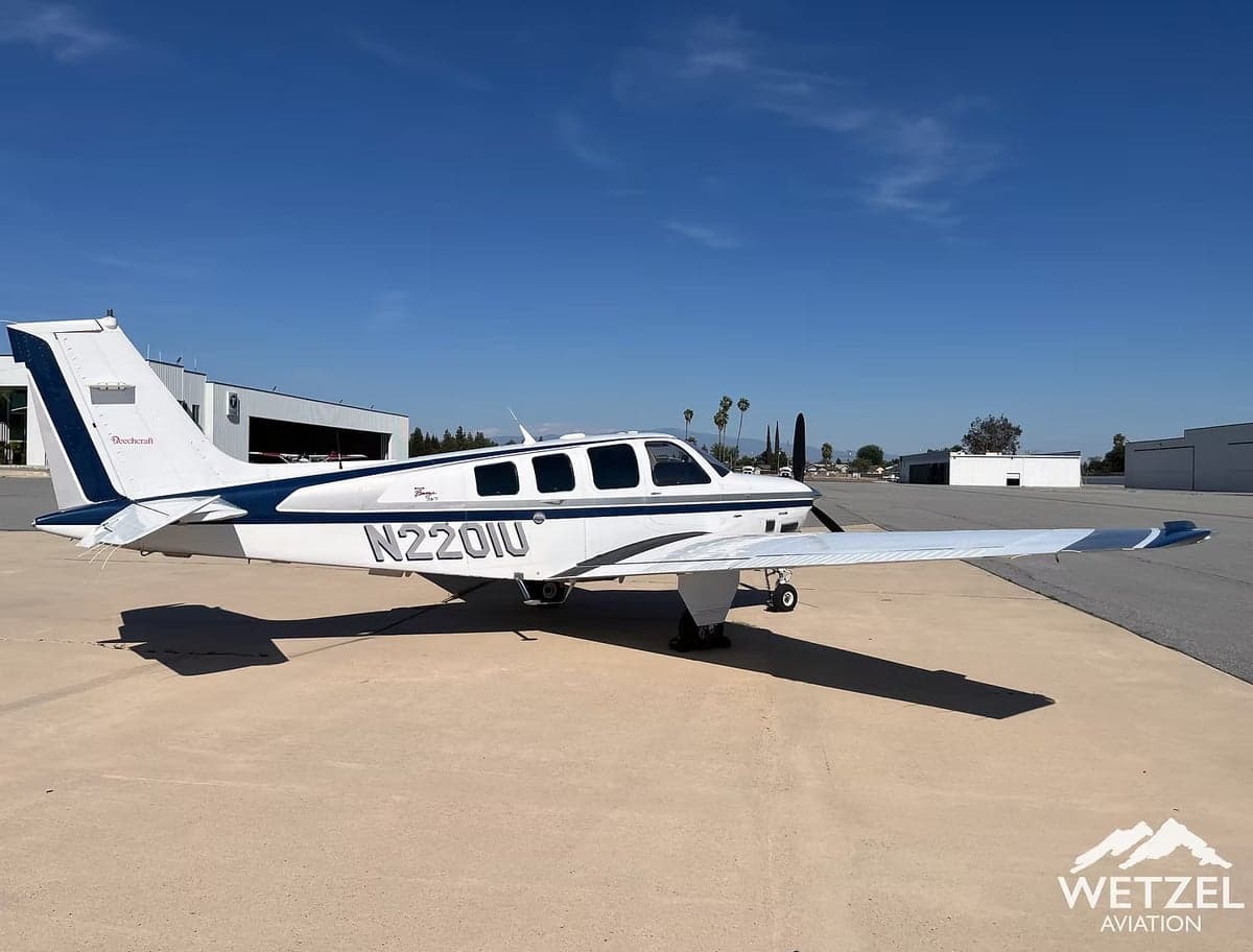 A Beechcraft Bonanza with tail number N220IU is parked on the tarmac in front of a hangar.