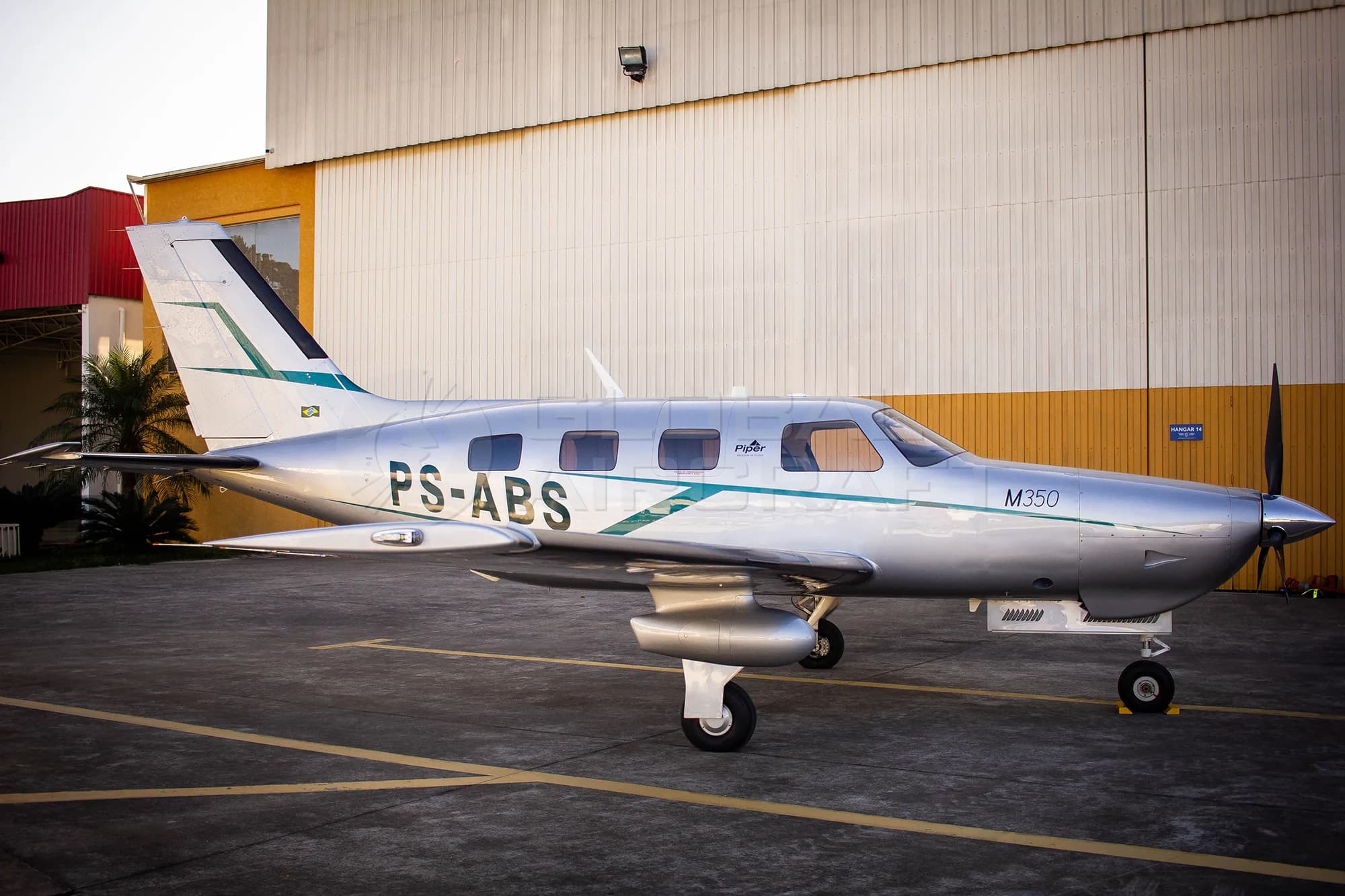 A silver Piper M350 aircraft with green and white stripes, tail number PS-ABS, parked on an tarmac in front of a hangar.