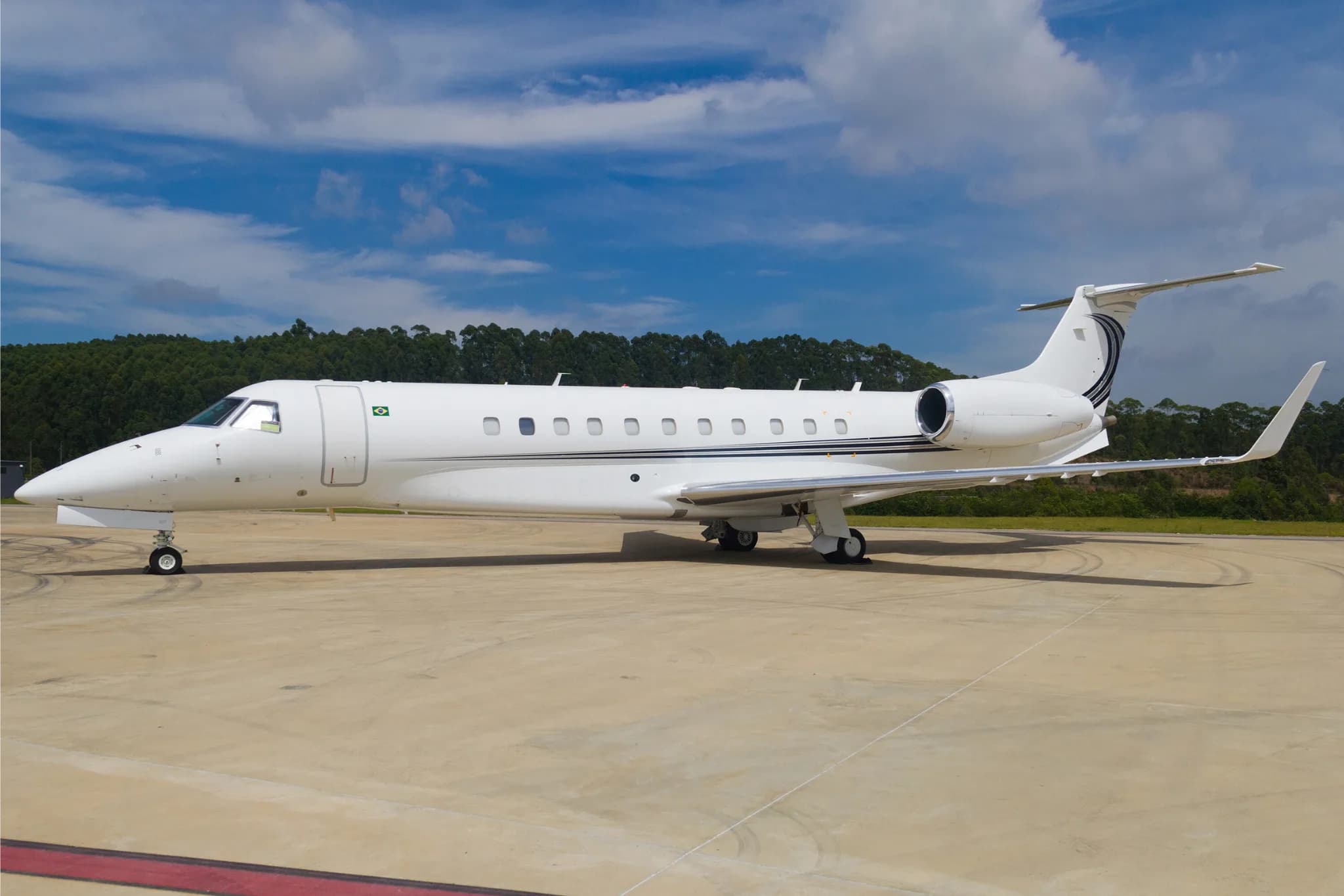 A white Embraer Legacy business jet with a black stripe on the tail, parked on a tarmac under a blue sky.
