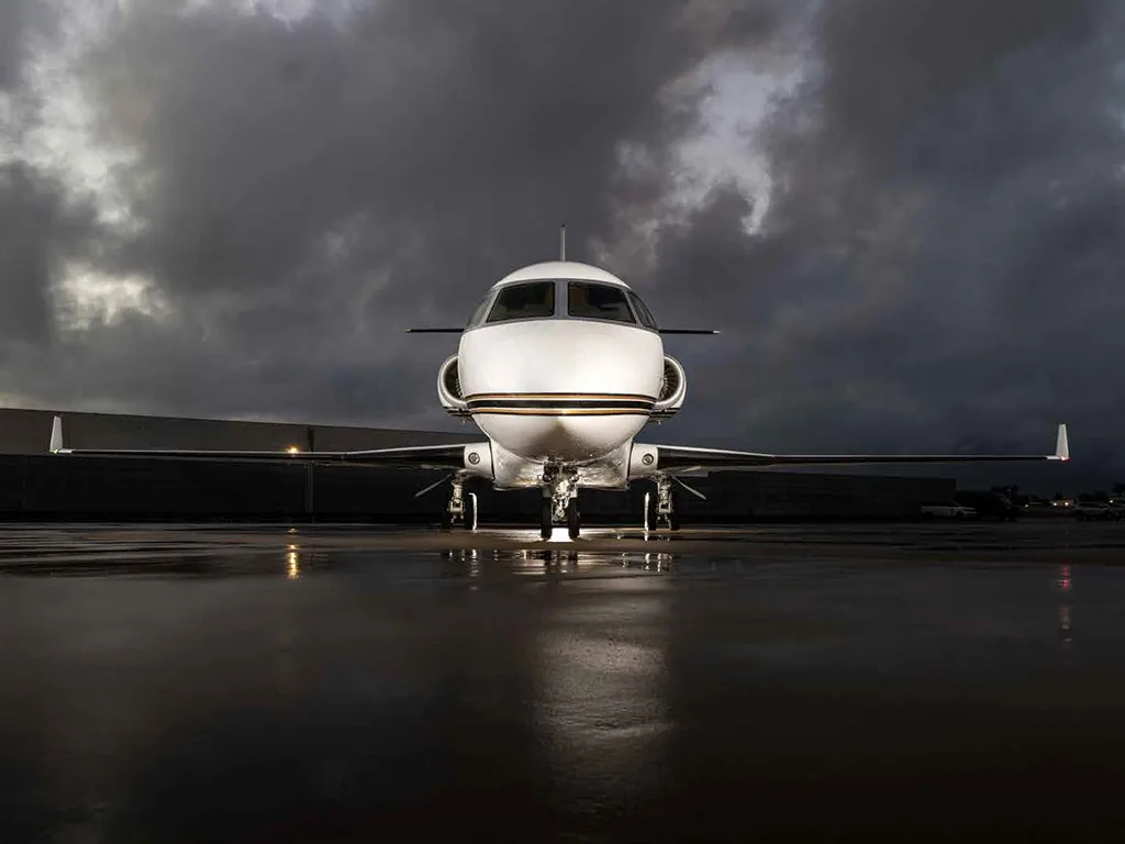 A head-on view of a white Gulfstream G450 business jet with a gold stripe on a wet tarmac at dusk.