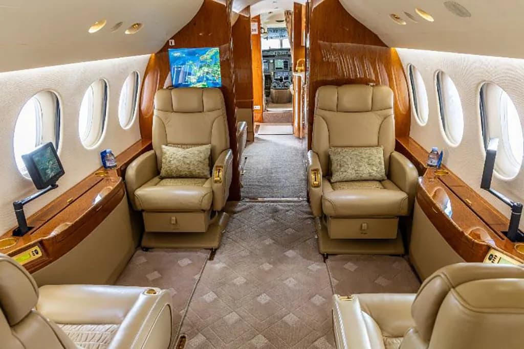 The private jet cabin interior with tan leather seats, wood paneling, and a view towards the cockpit.