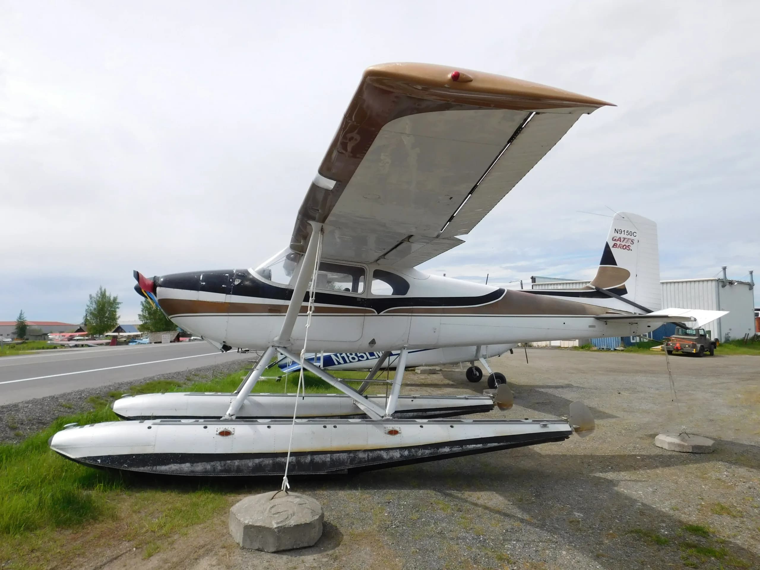 A white and brown Cessna 185 Skywagon floatplane is parked on a gravel lot next to a road.