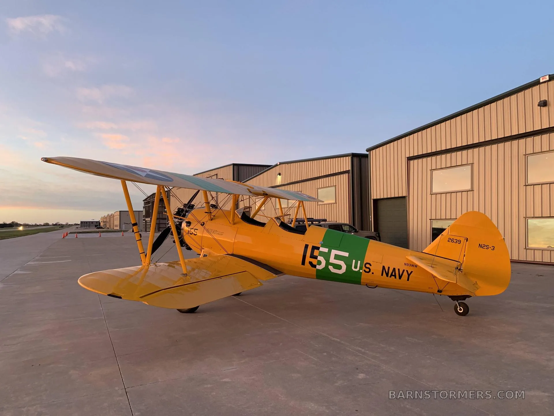 A yellow biplane with U.S. Navy markings and the number 155, parked on an airfield apron in front of hangars at dusk.