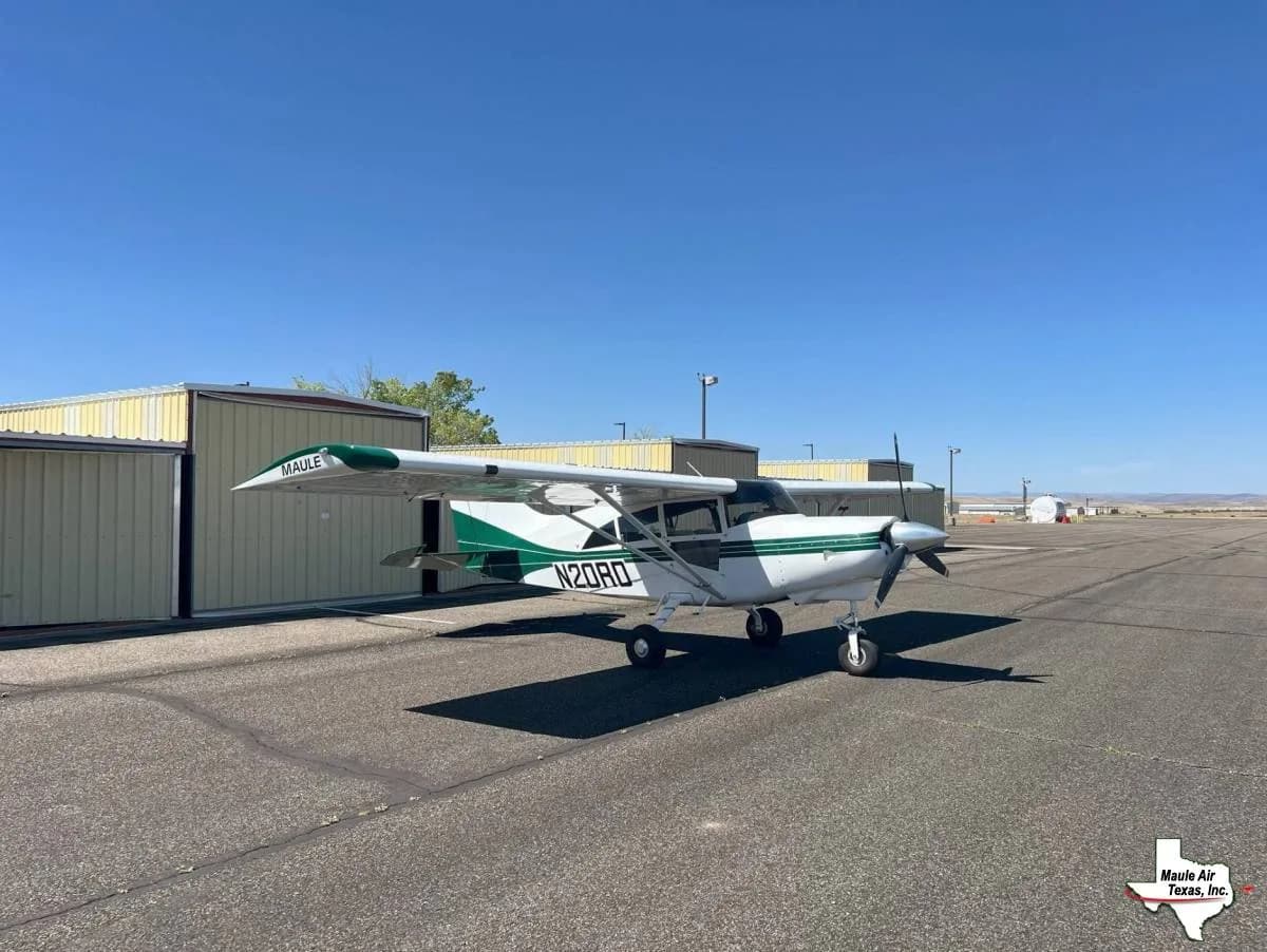 A white and green Maule aircraft, registration N20RD, parked on a tarmac next to hangars under a clear blue sky.