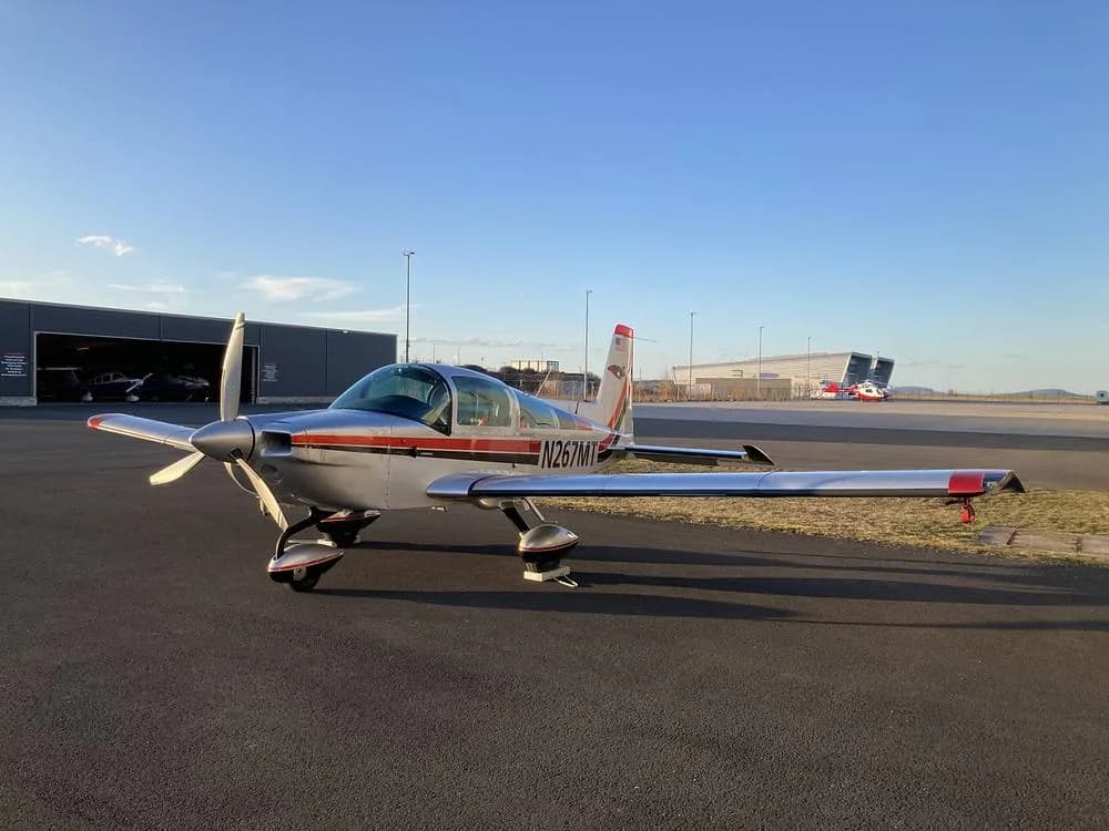 The Grumman American AA-5B Tiger aircraft, N267MT, parked on an airfield with hangars in the background under a clear sky.