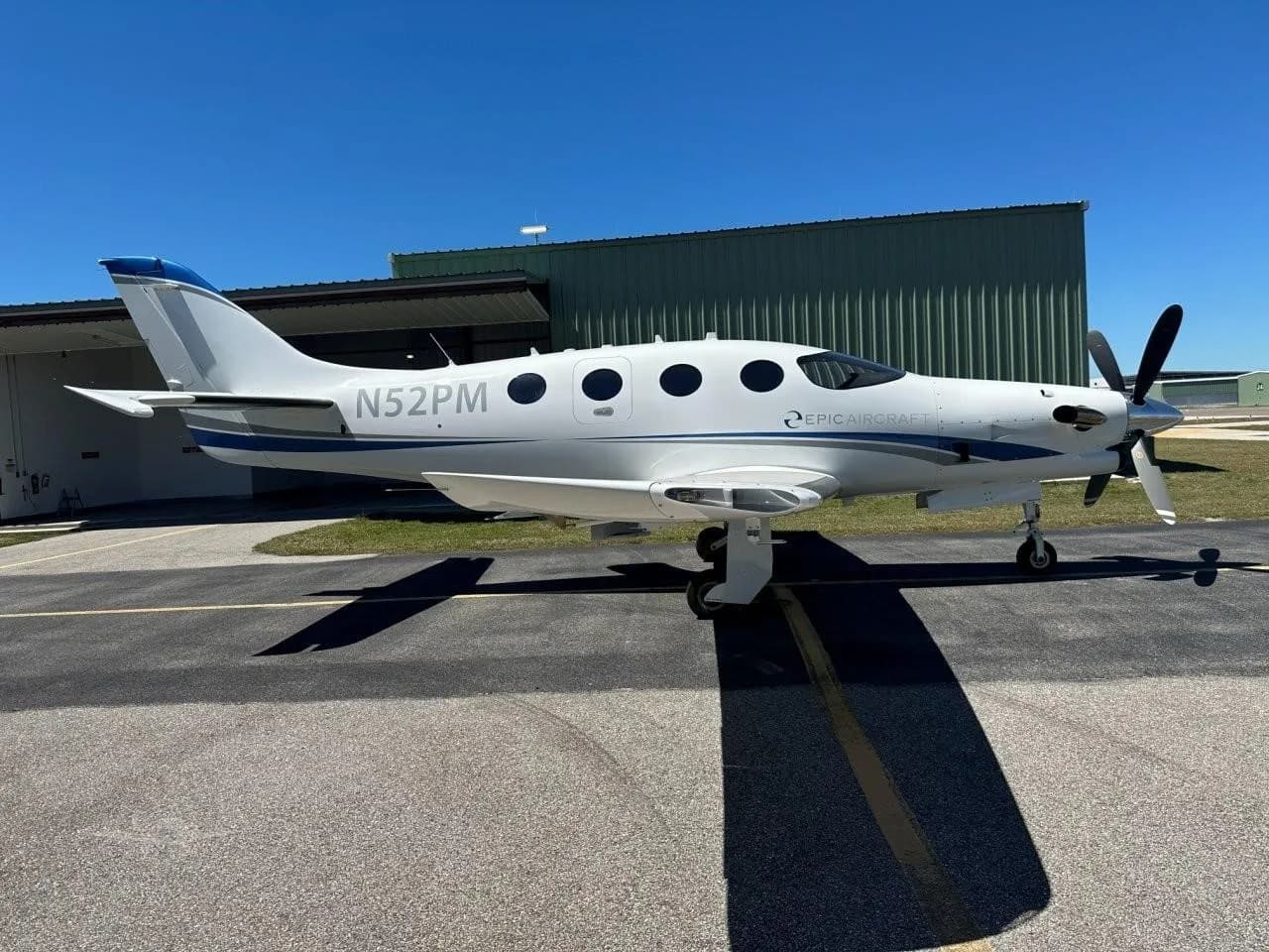 A white Epic E1000 turboprop aircraft with blue accents and registration N52PM, parked on an airfield in front of a green hangar.