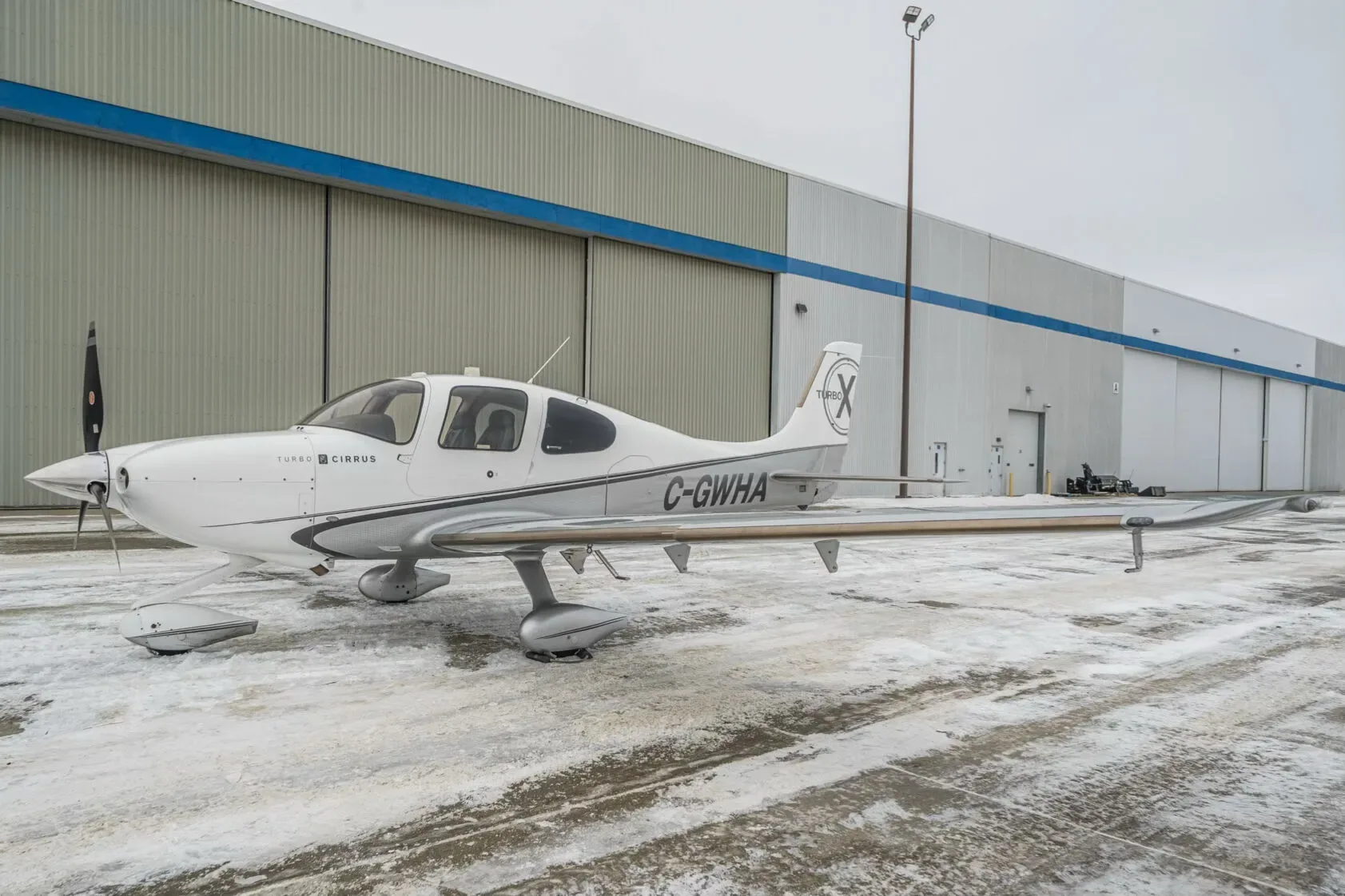 A white and grey Cirrus SR22T aircraft (C-GWHA) with a 'Turbo X' logo on the tail, parked on a snowy ground next to a large hangar.