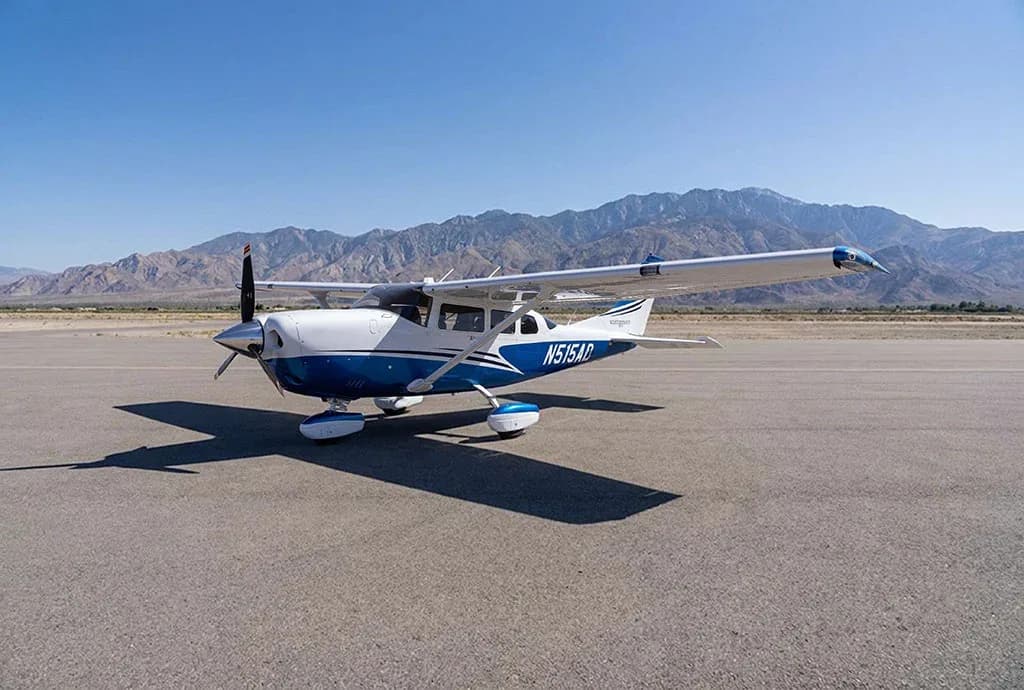 A white and blue Cessna Stationair N515AD parked on an airfield with mountains in the background.