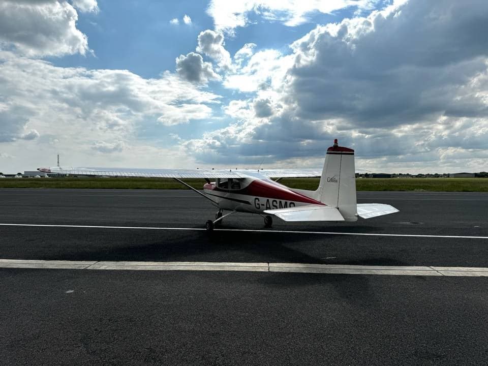 A white and red Cessna 150 G-ASMS parked on the tarmac, viewed from the rear-right, under a bright cloudy sky.