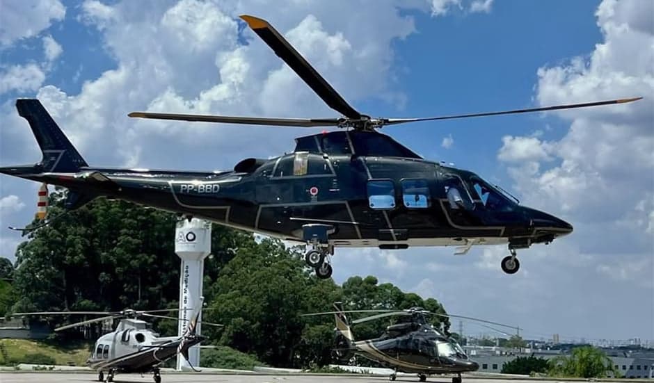 A black helicopter with registration PP-BBD is shown in flight with two other helicopters on the ground.