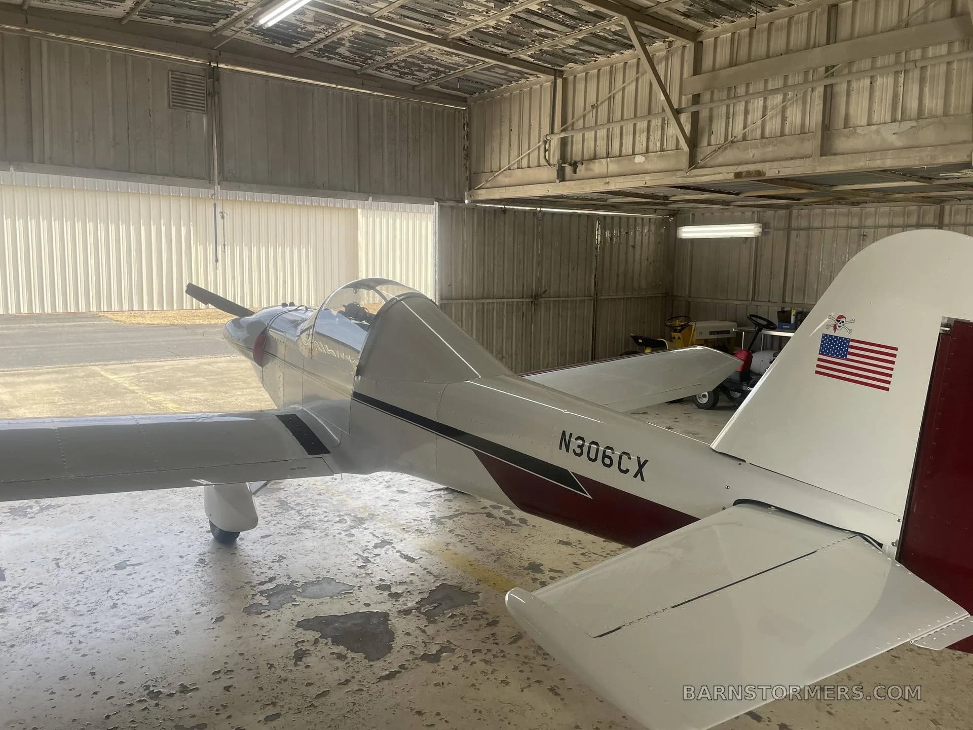 A rear-quarter view of a white and maroon experimental aircraft with an American flag on the tail, parked in a hangar.