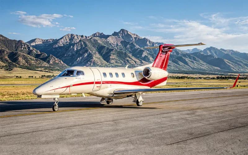 A white Learjet 75 business jet with red stripes on the fuselage and tail, parked on a runway with mountains in the background.