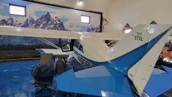 The rear-right side of a white and blue Maule aircraft, N1008W, parked in a hangar with a mountain mural.