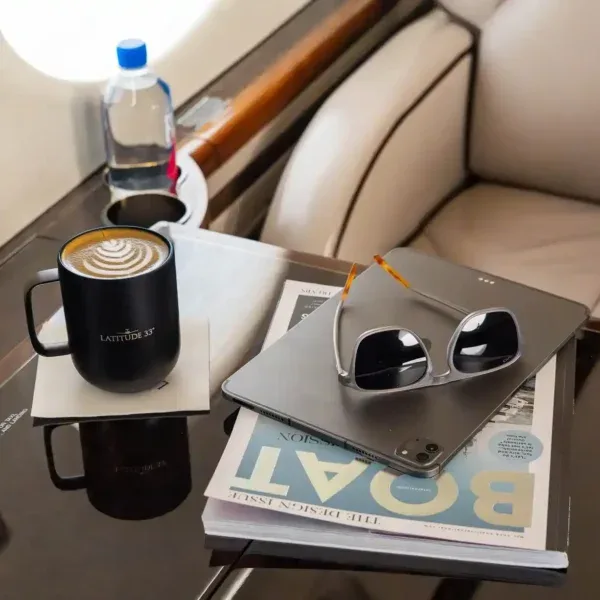 A close-up of a coffee cup, sunglasses, a tablet, and a magazine on a polished table inside the aircraft cabin.