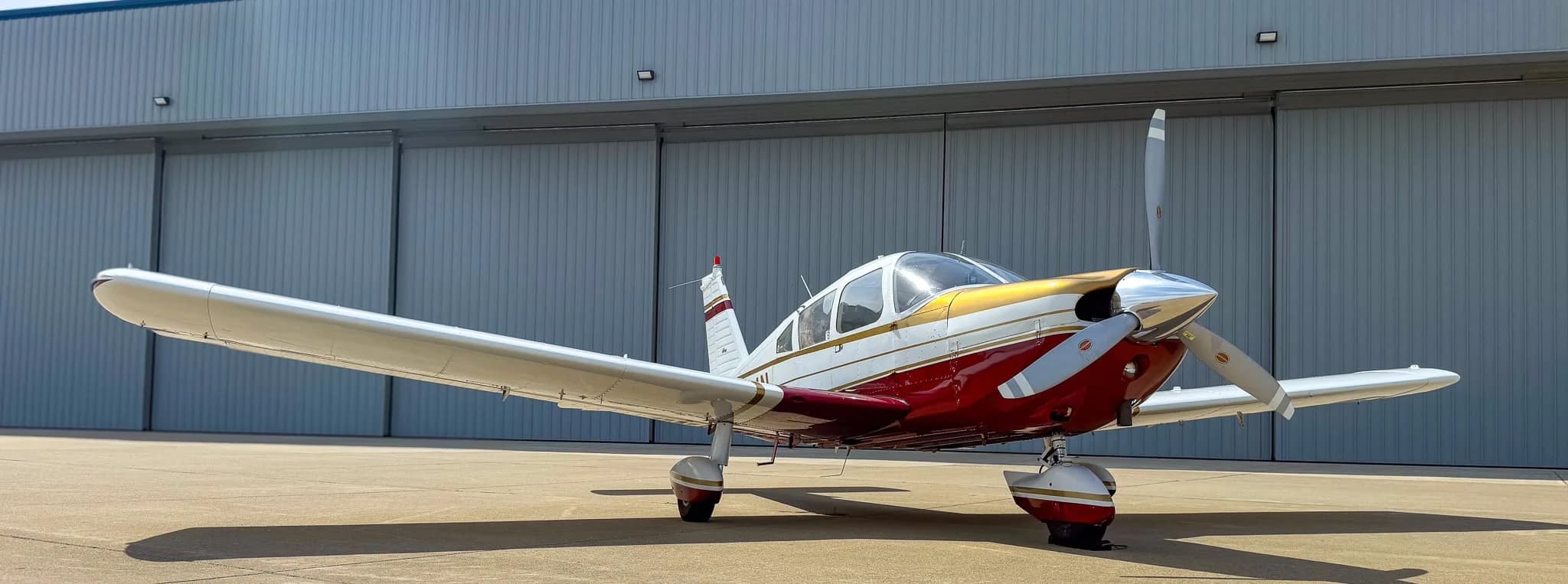 A white, red, and gold Piper Cherokee aircraft (N8534N) parked on an airfield with a hangar in the background.