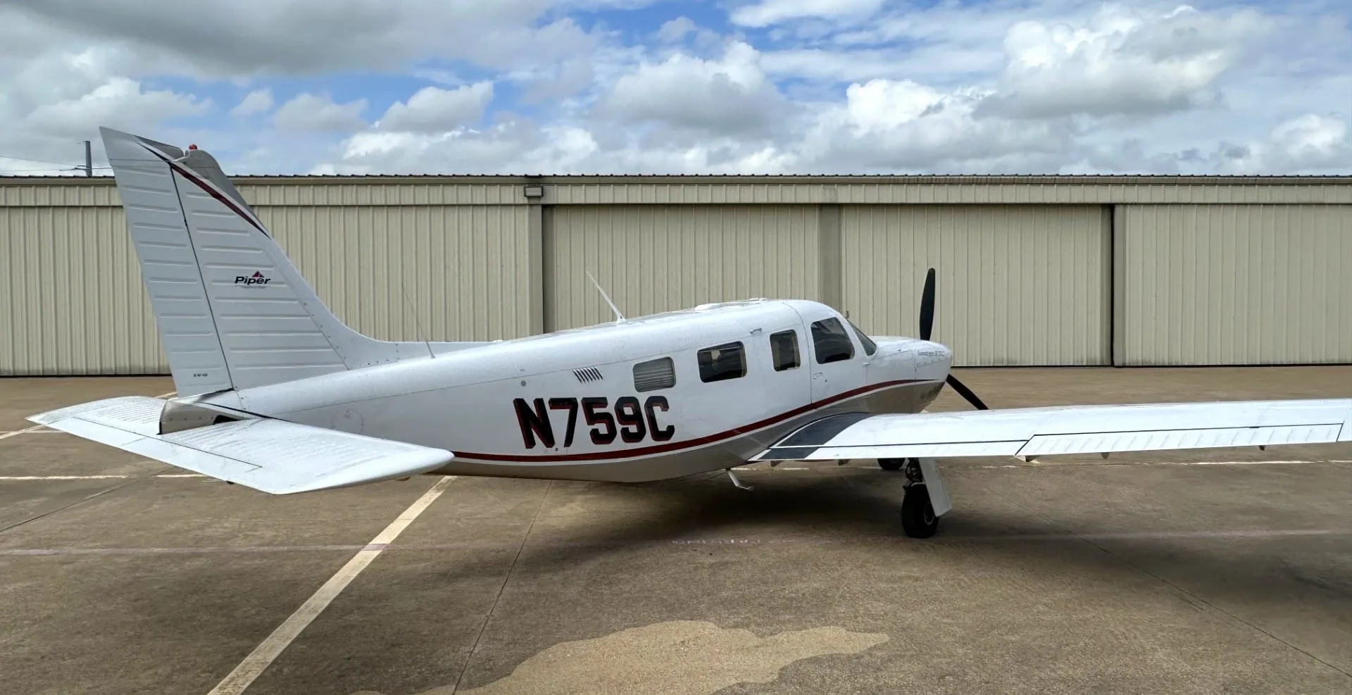 A white and tan Piper Saratoga II TC aircraft (N759C) parked on an asphalt ramp in front of hangars under a cloudy sky.