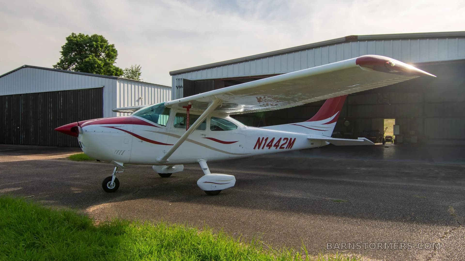 A white single-engine Cessna aircraft with red and gold stripes is parked on an asphalt ramp, viewed from the front-left.