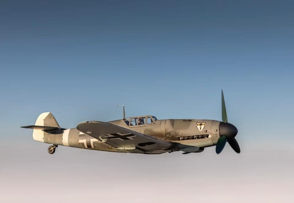 A gray Messerschmitt Bf 109 aircraft in flight against a blue sky.