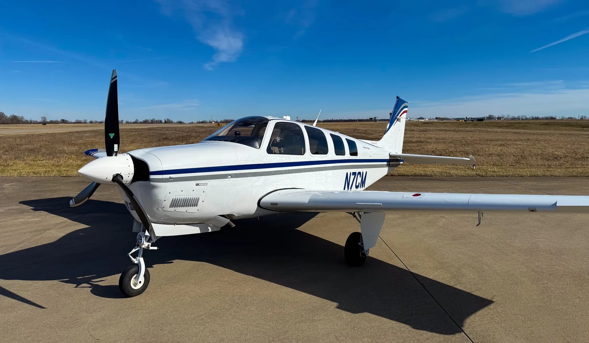 A white Beechcraft Bonanza G36 with blue and grey stripes, registration N7CM, parked on an airfield under a clear blue sky.