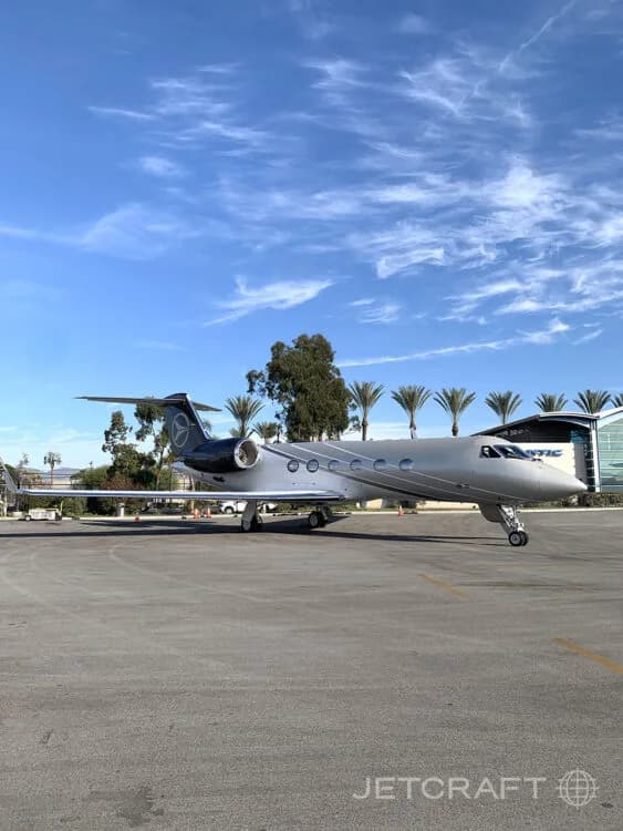 A silver and dark blue Gulfstream G450 business jet parked on a tarmac with palm trees and a building in the background.