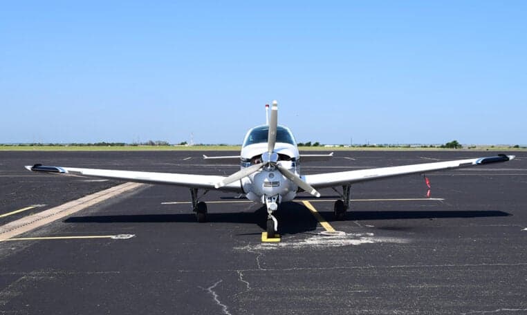 A head-on view of a white and blue Beechcraft Baron aircraft with its propeller centered on an asphalt tarmac.