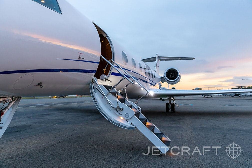 The open main entry door and illuminated airstairs of a white Gulfstream G550 business jet on the tarmac at dusk.