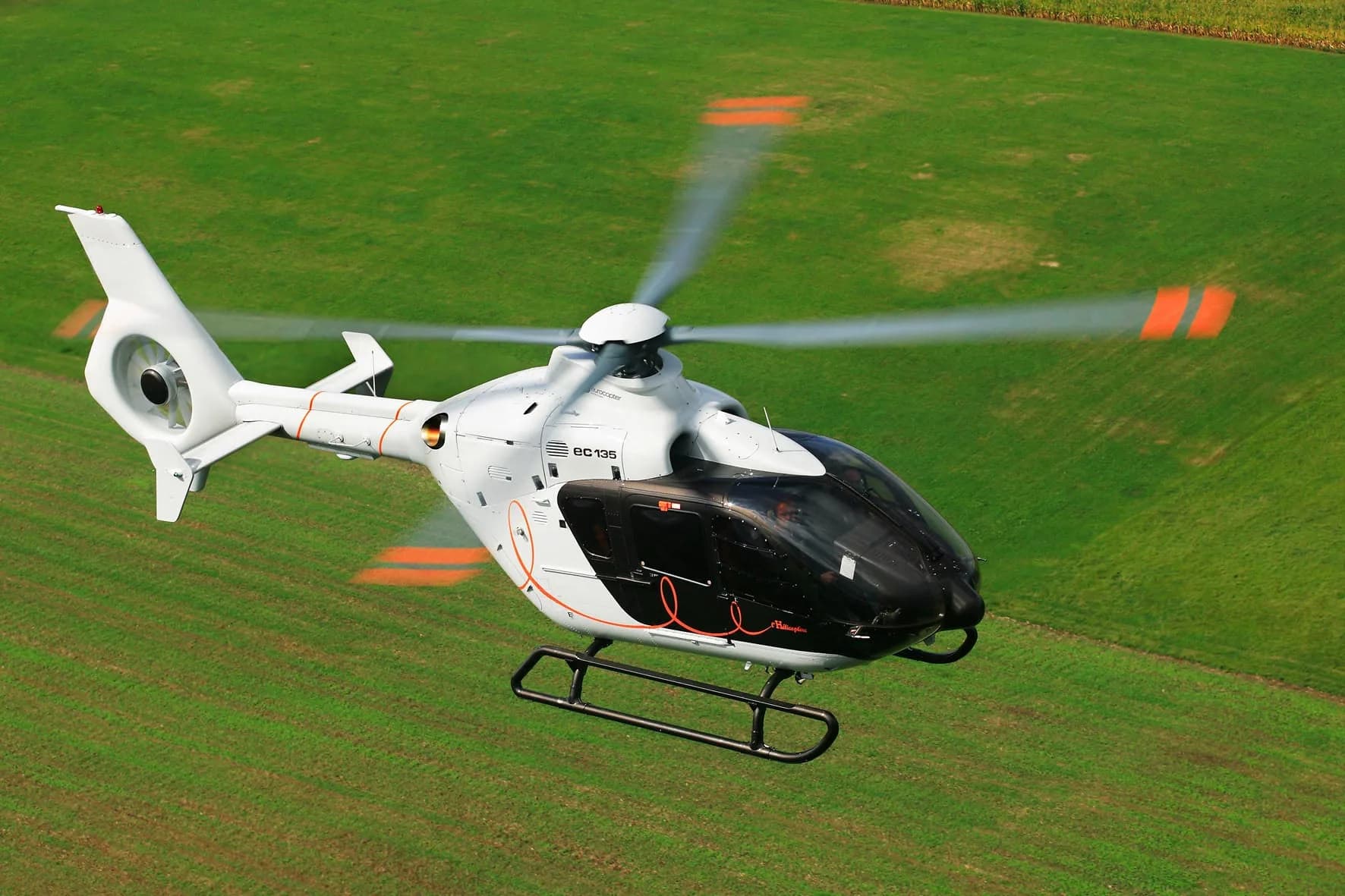 A white and black Airbus EC135 helicopter in flight over a green field.