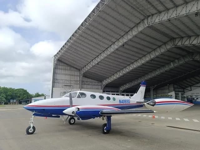 A three-quarter front view of a white and blue Cessna 340 twin-engine aircraft with red stripes parked next to a hangar.