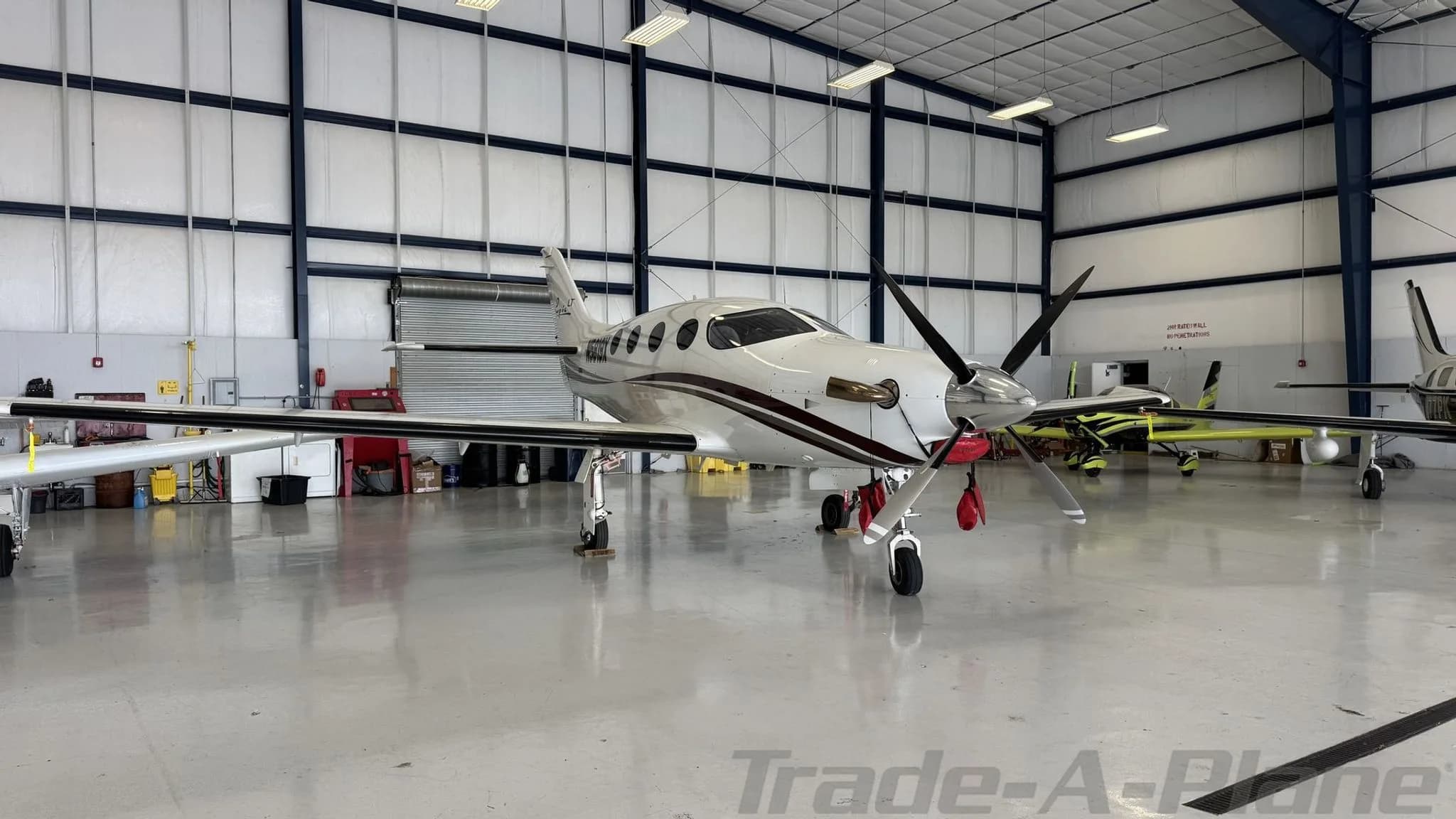 Wide view of a white Epic LT aircraft with a dark red stripe, parked in a hangar with other aircraft.