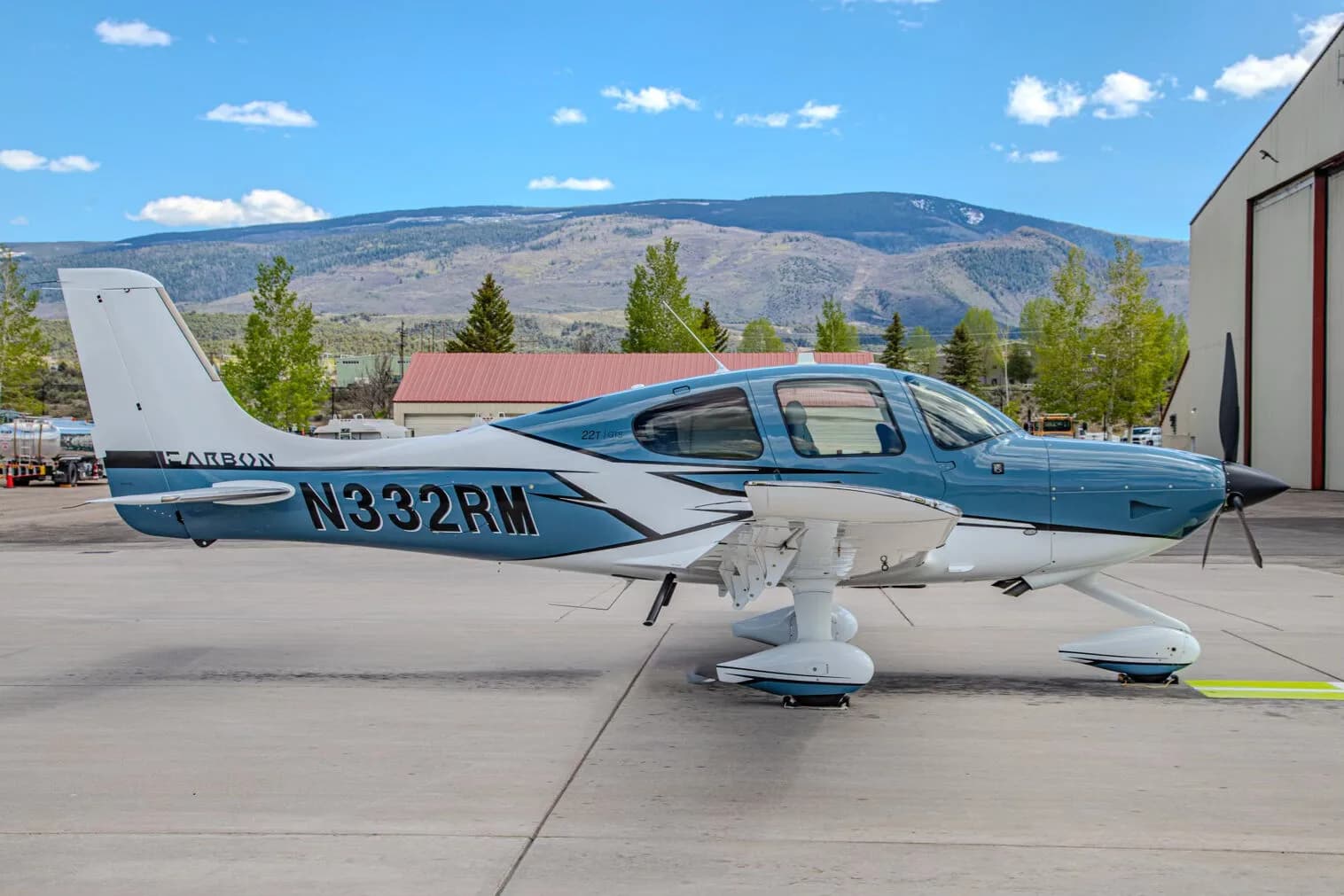 A blue and white Cirrus SR22T GTS aircraft, registration N332RM, parked on an airport tarmac next to a hangar with mountains in the background.