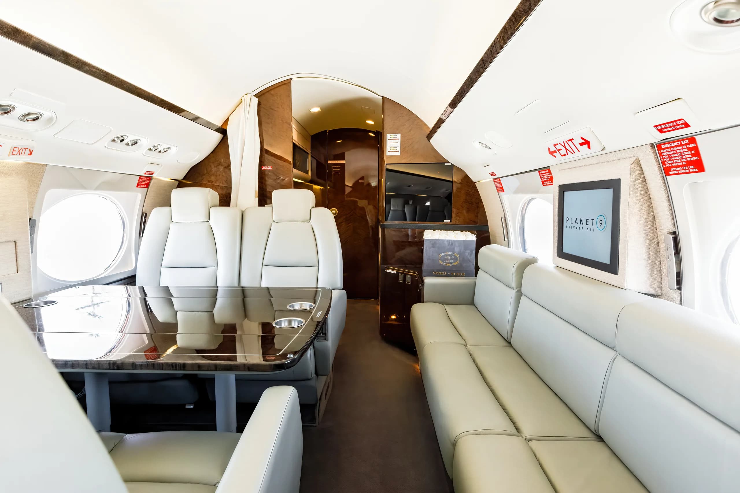 The luxurious interior cabin of a private jet features light gray leather seats, a dark polished table, and a screen displaying "PLANET 9 PRIVATE AIR".