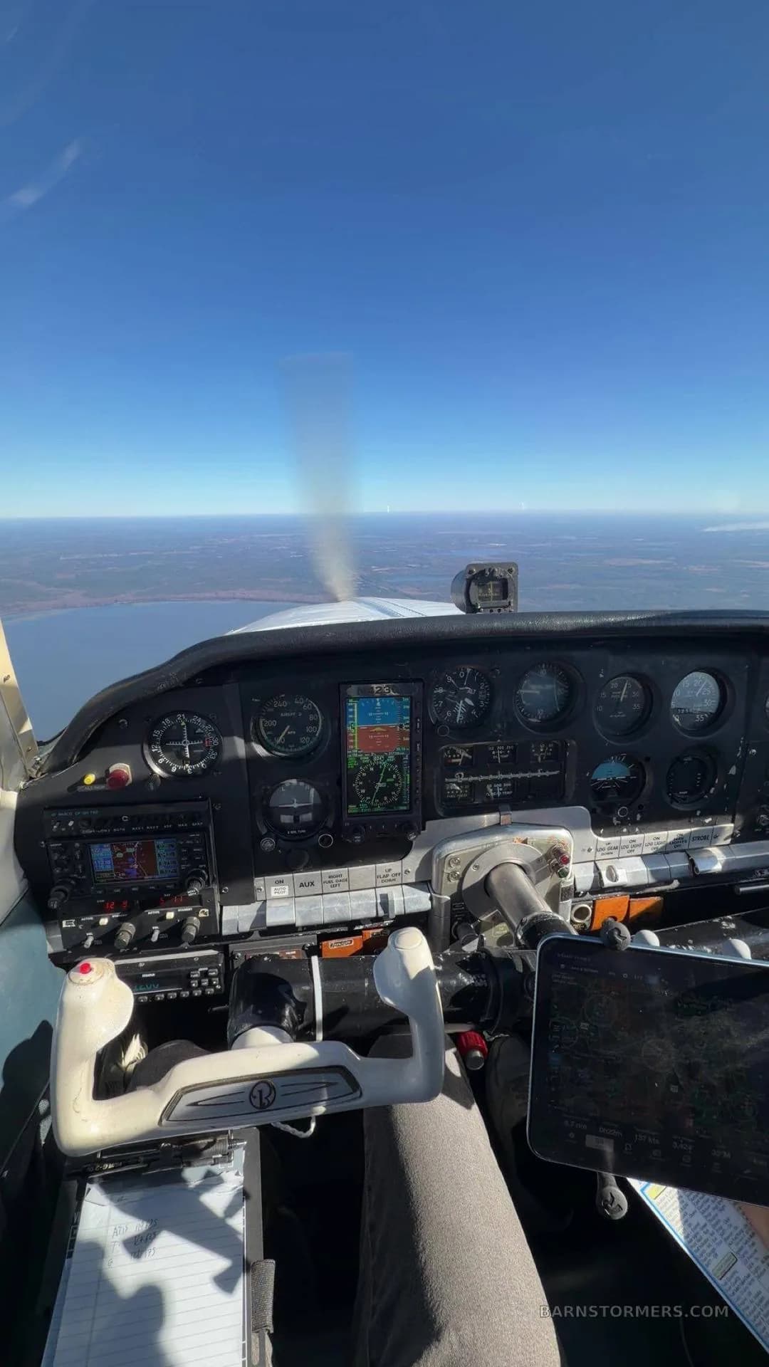 The cockpit of a small aircraft in flight, showing the instrument panel, controls, and a view of land and water below.