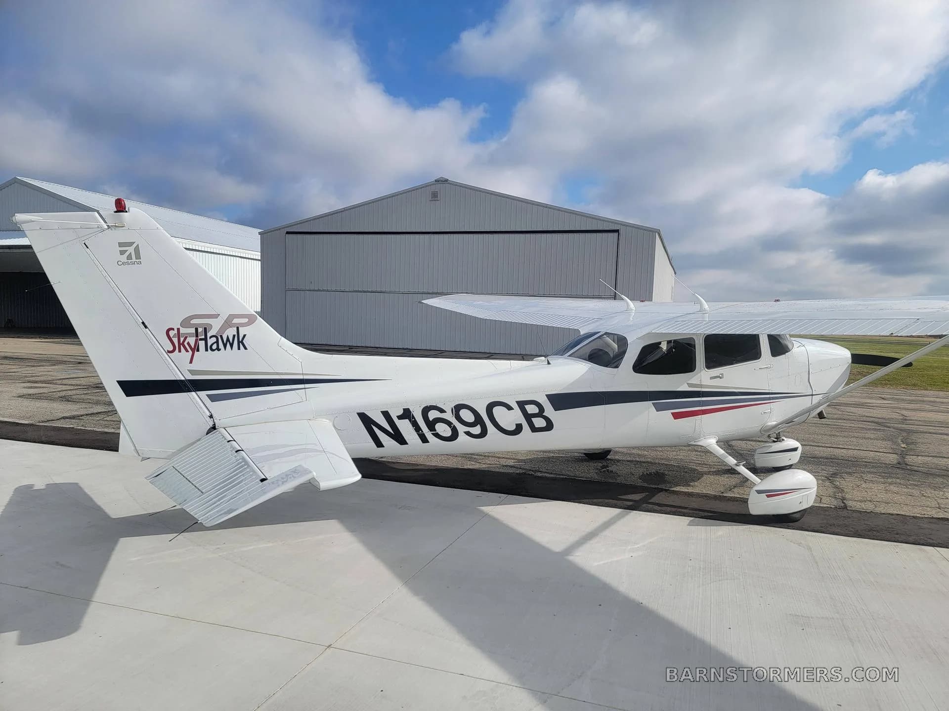 A white Cessna Skyhawk N169CB with black and red stripes is viewed from the rear-right, parked on an apron near hangars.