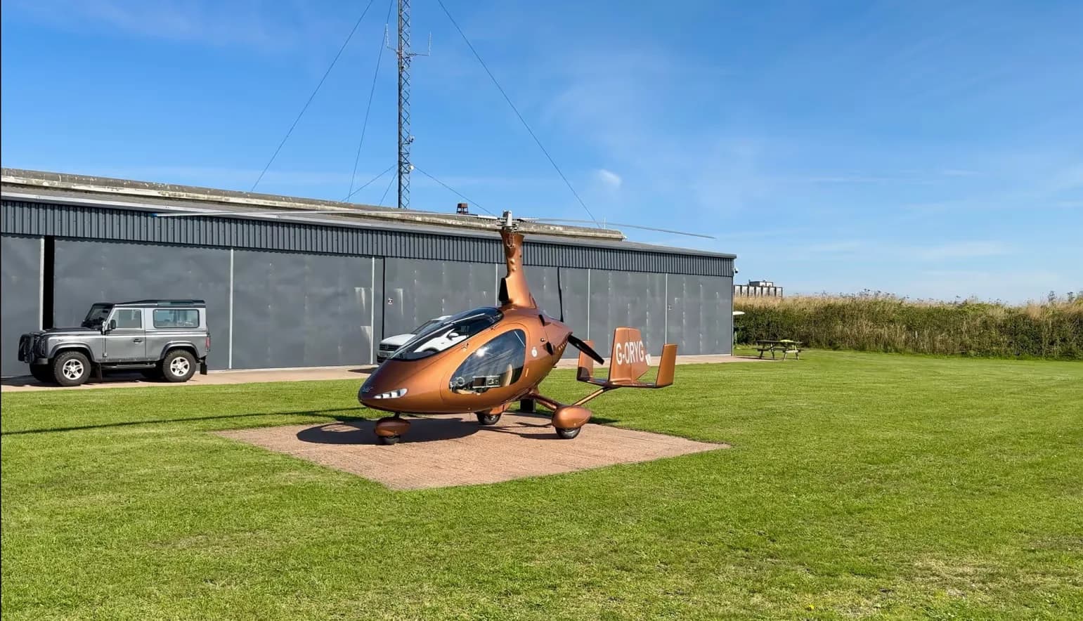 A brown gyrocopter, G-ORYG, parked on a concrete pad in front of a hangar with a grey SUV.