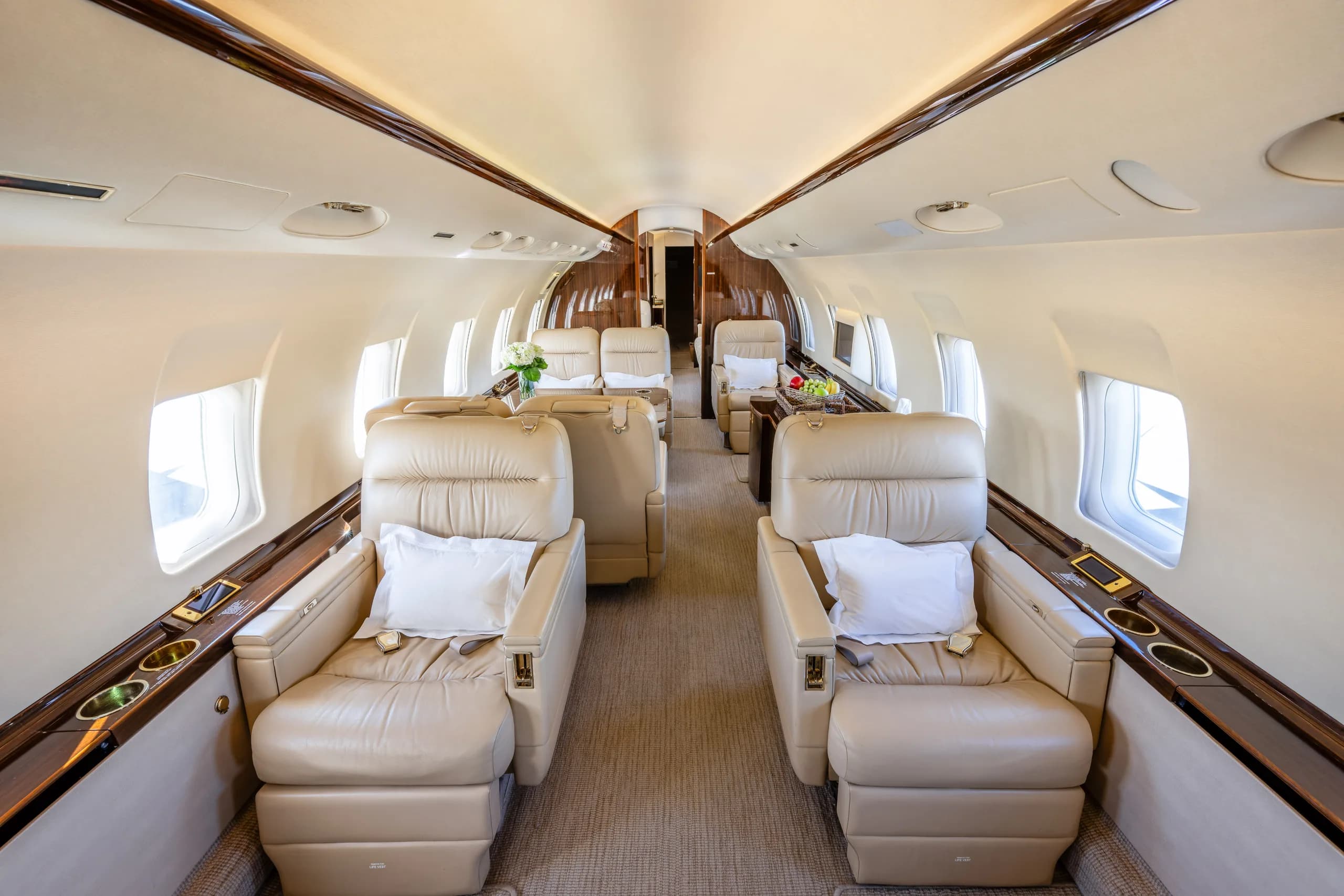 The luxurious cabin interior of a private jet, featuring two rows of plush beige leather seats facing each other, with white pillows and dark wood accents.