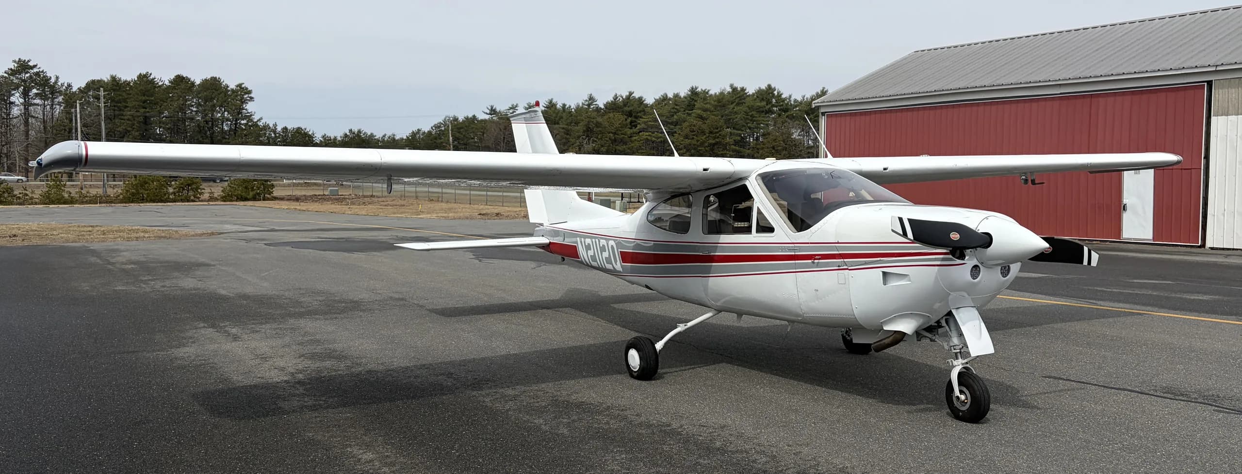 A white Cessna Cardinal RG with red and grey stripes parked on an asphalt ramp in front of a hangar.