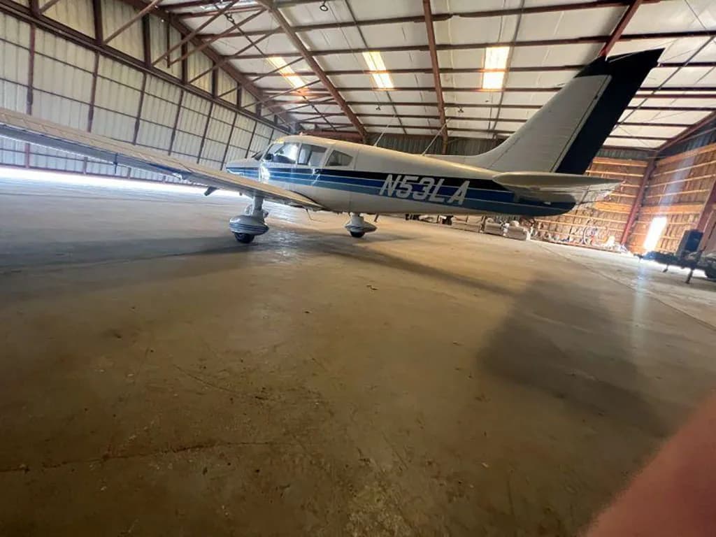 A low-angle rear-left view of the white Piper Cherokee aircraft N53LA with blue stripes, parked in a hangar.