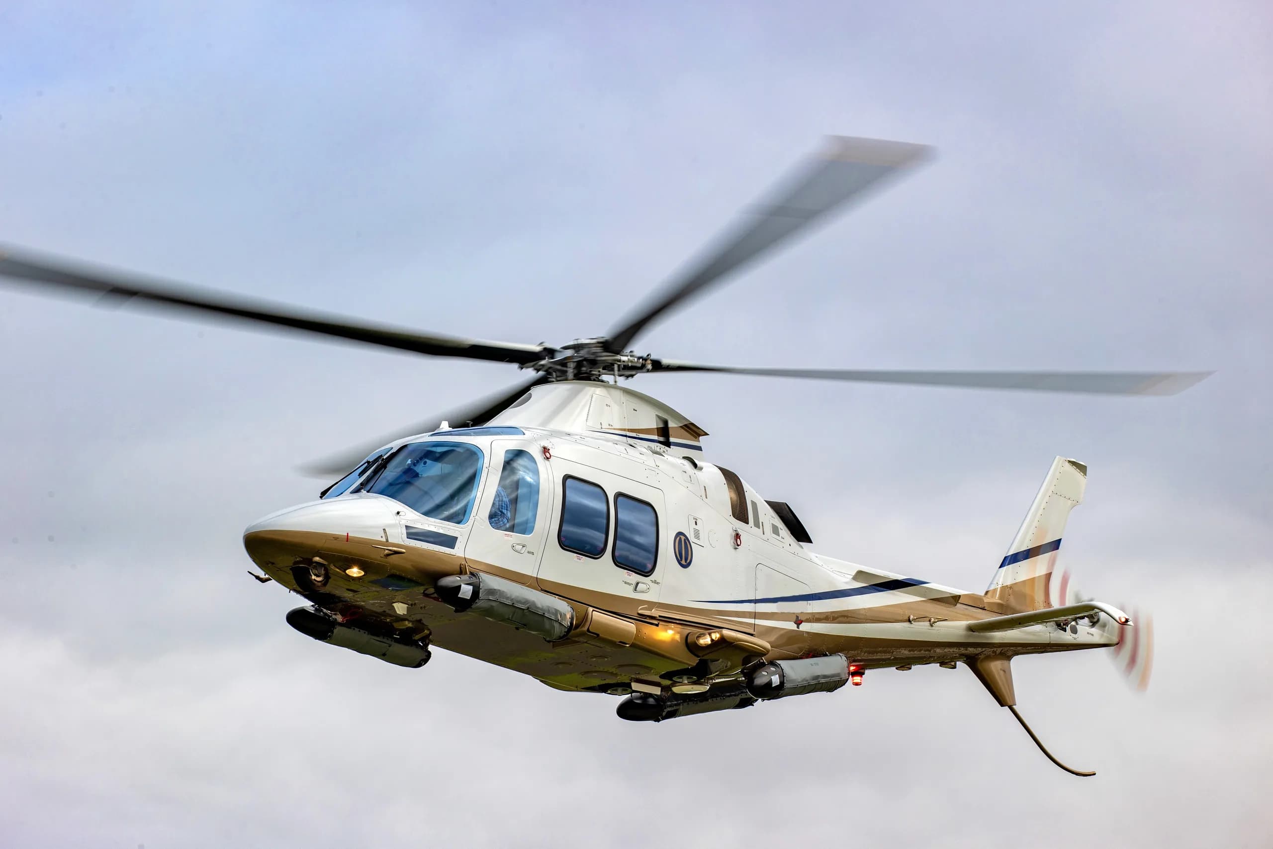 A white and gold helicopter in flight against a cloudy sky.