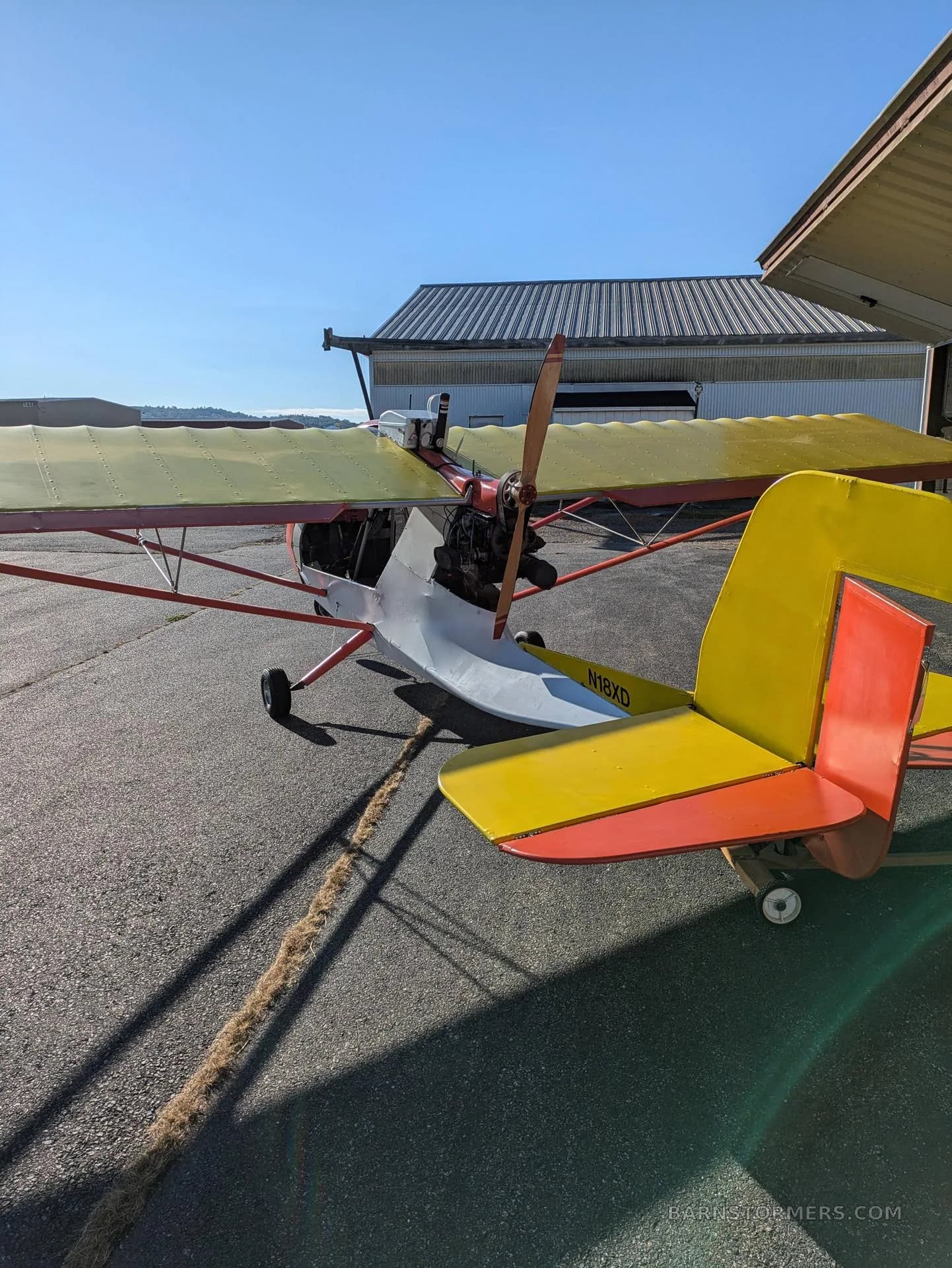 A rear-quarter view of a red and yellow ultralight aircraft with a wooden propeller and registration N18XD.
