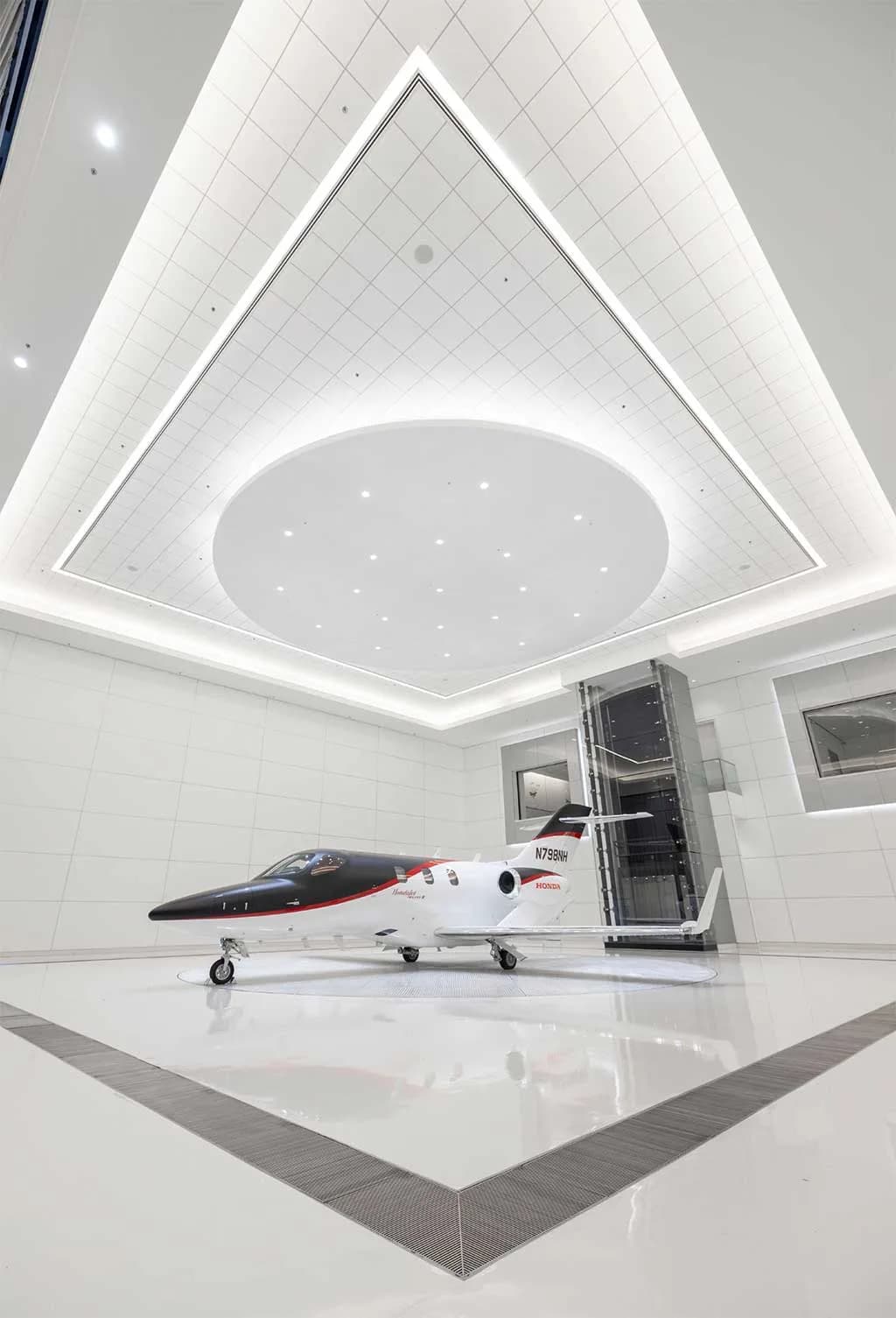 A black and white HondaJet Elite II (N798NH) with red accents is viewed from a high angle in a modern white showroom with a distinctive ceiling design.