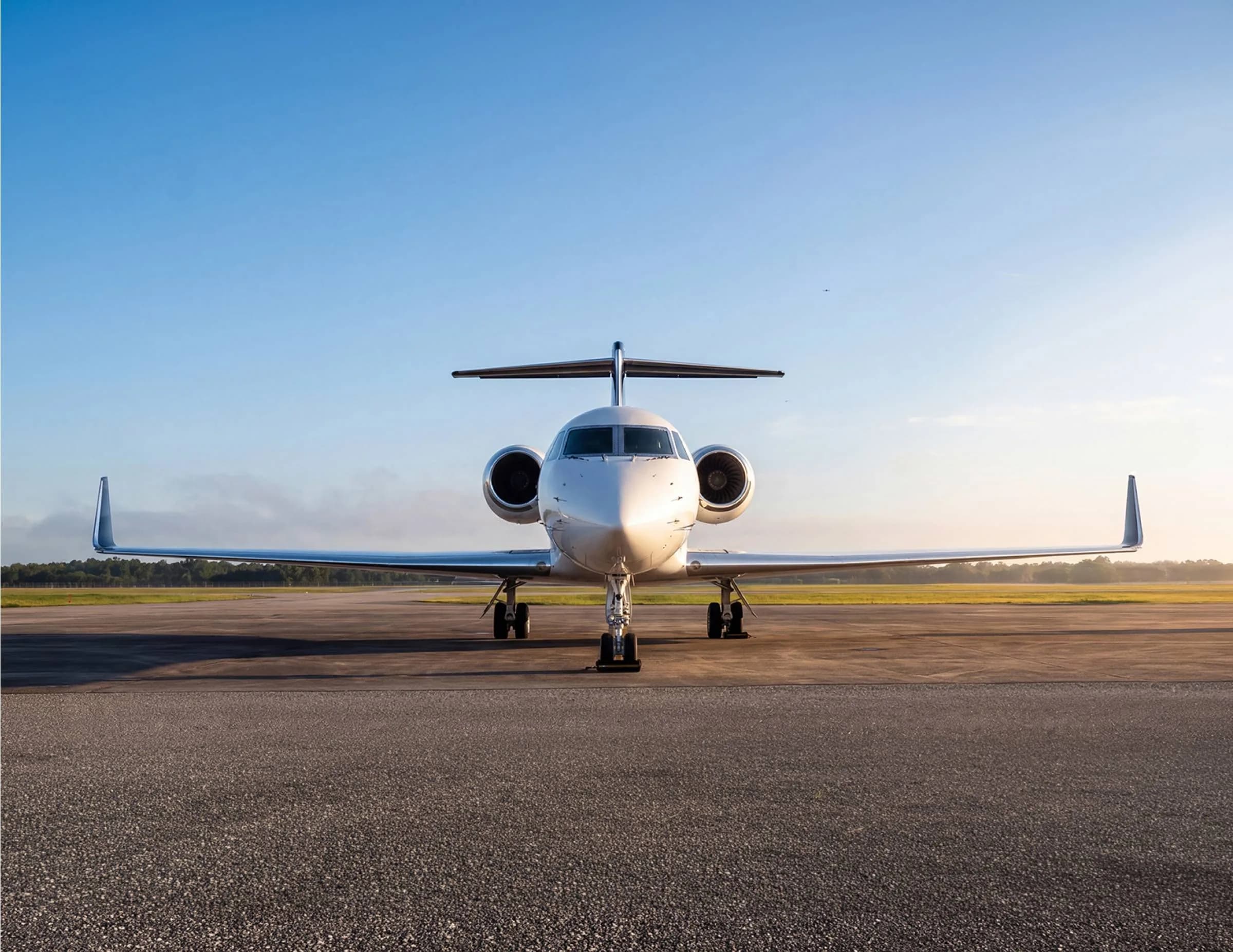 The front view of a white business jet with a dark blue tail parked on an airfield at sunrise or sunset.