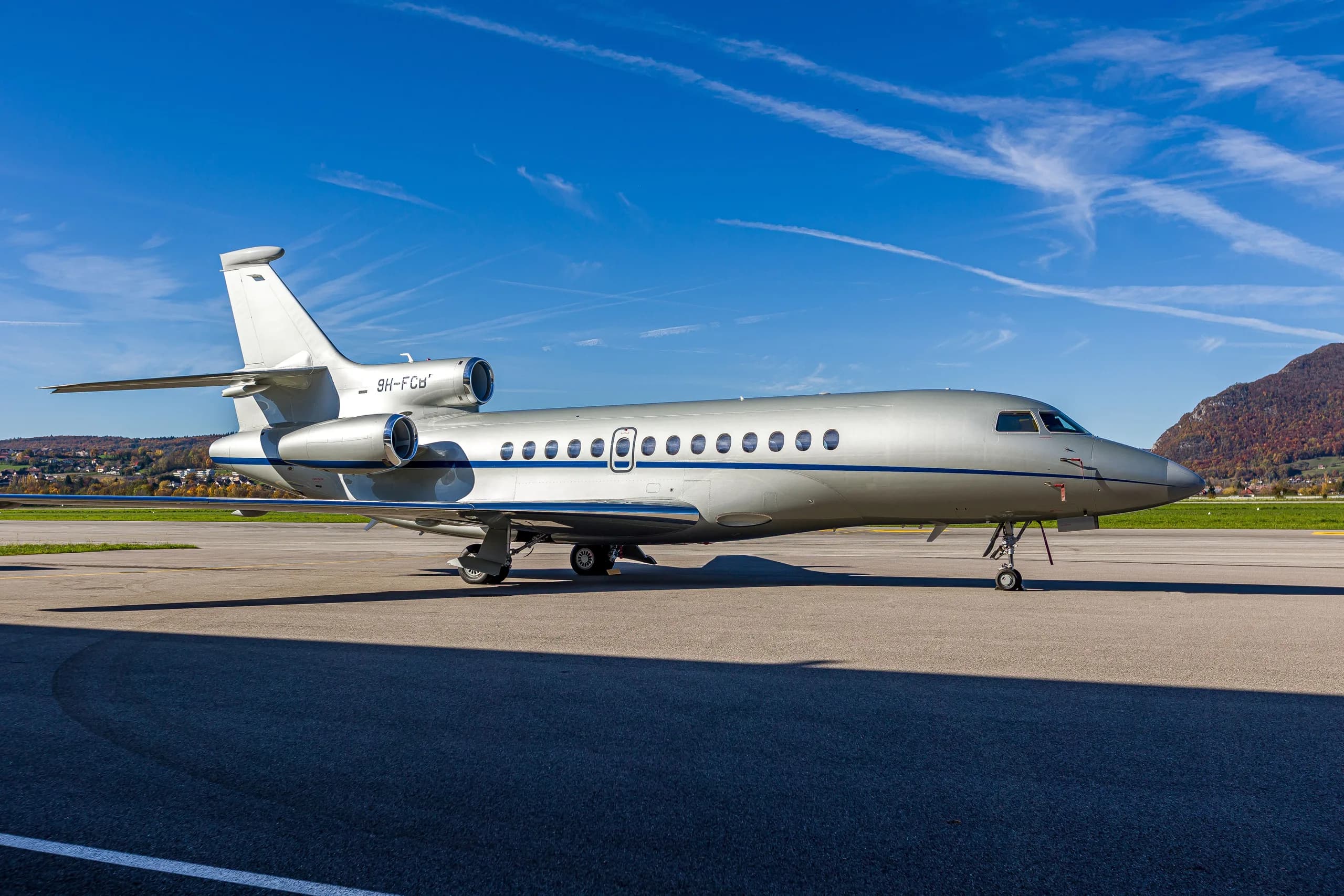 A silver Dassault Falcon 900EX business jet with blue stripes parked on an airfield under a clear blue sky.