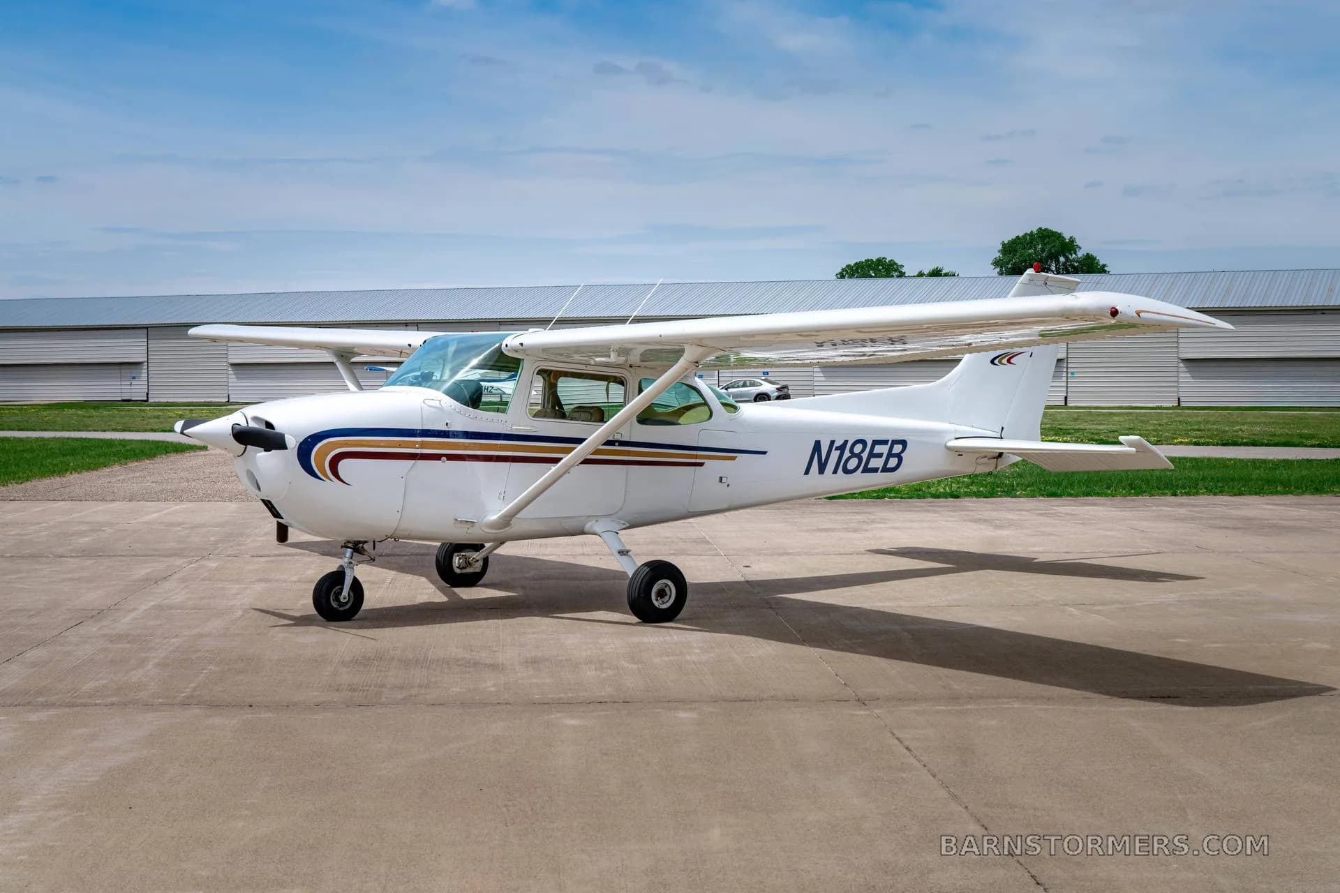 A white Cessna 172 (N18EB) with blue and gold stripes is parked on a tarmac, viewed from the front-right.