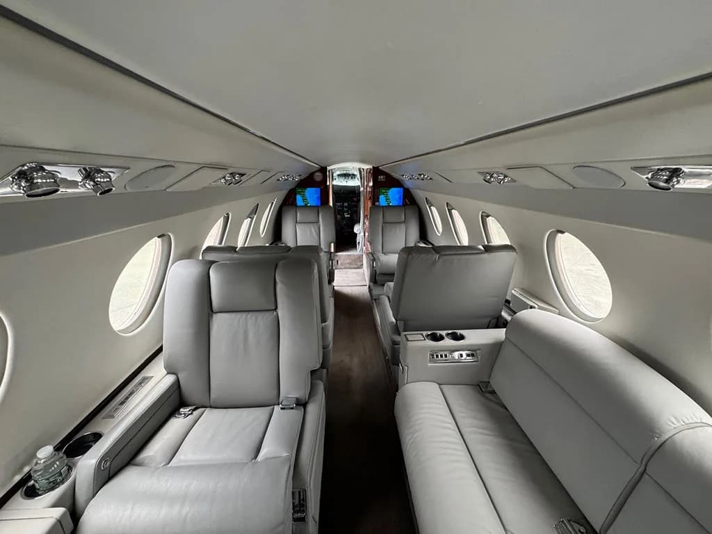 View down the aisle of a business jet cabin with light gray leather seats and wood accents, looking towards the cockpit.