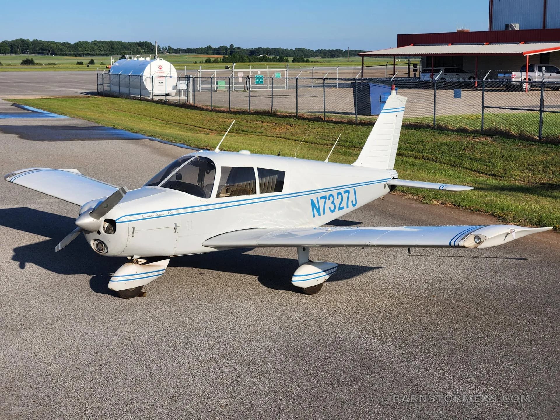 A white Piper Cherokee aircraft with blue stripes, registration N7327J, parked on an asphalt ramp at an airfield.