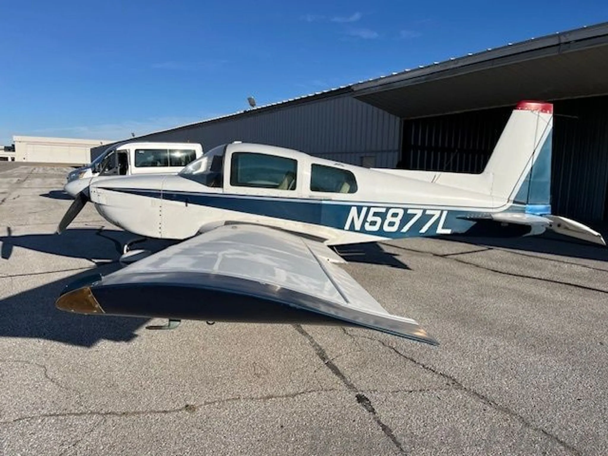 A white and blue Grumman AA-5B Tiger aircraft (N5877L) parked on a tarmac next to a hangar.
